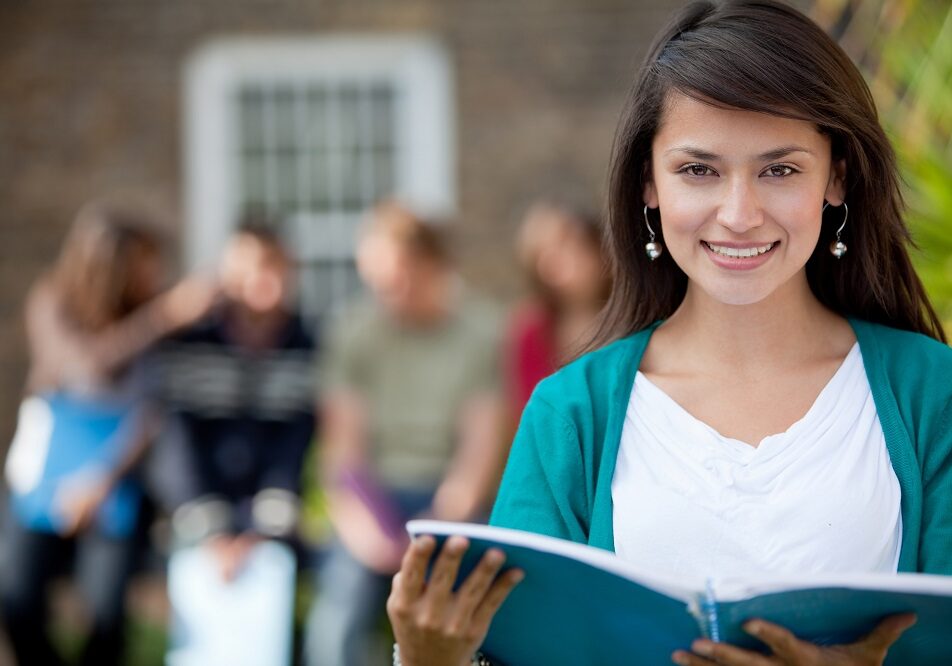 College aged female on a college campus reading a notebook and smiling. Peers behind her talking.